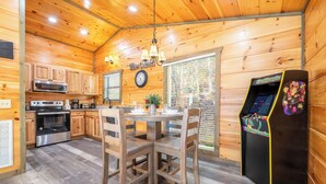 Interior de una cabaña de madera con cocina, comedor y máquina de videojuegos.
