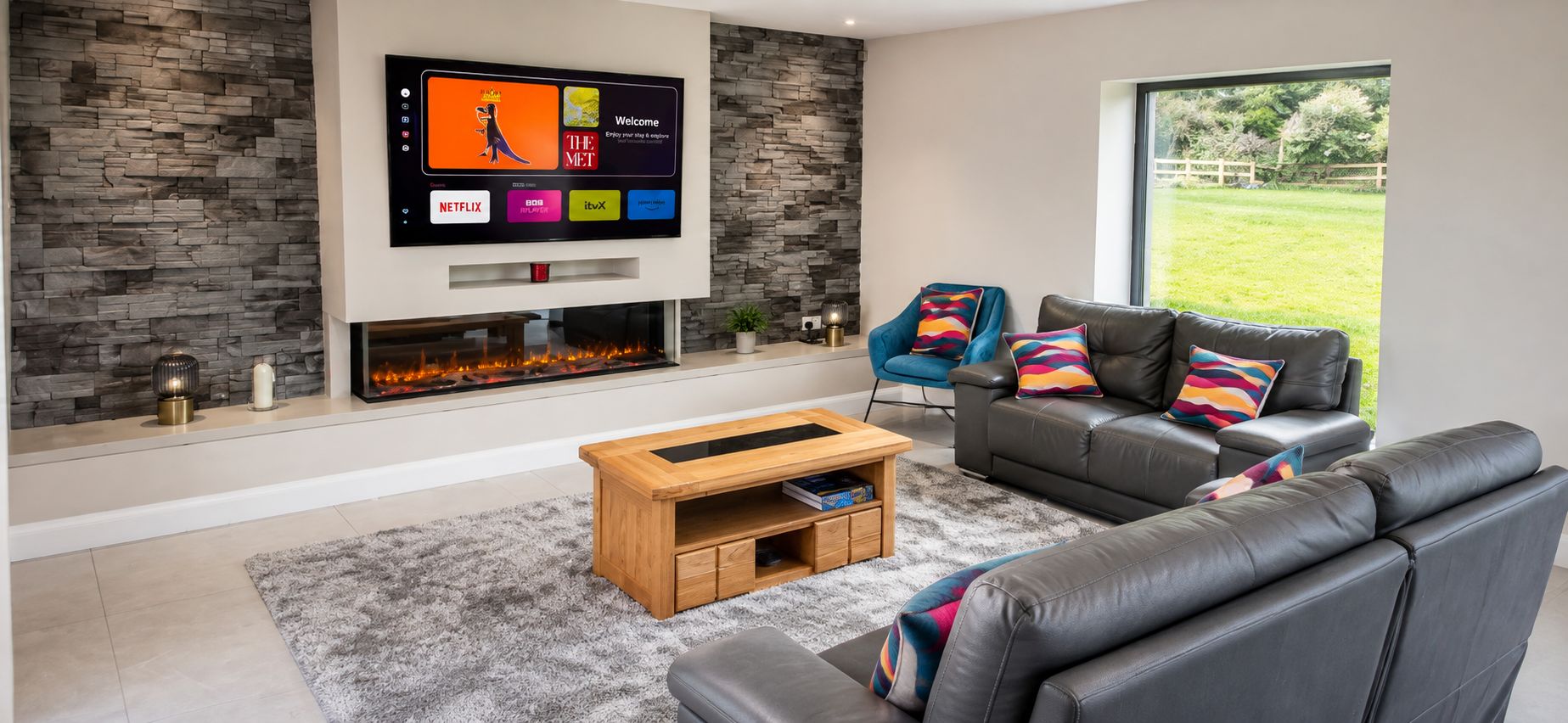 A modern living room with a stone wall, a flat-screen TV mounted above a fireplace, a grey leather sofa, a blue armchair, and a wooden coffee table.