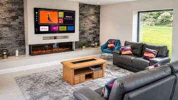 A modern living room with a stone wall, a flat-screen TV mounted above a fireplace, a grey leather sofa, a blue armchair, and a wooden coffee table.