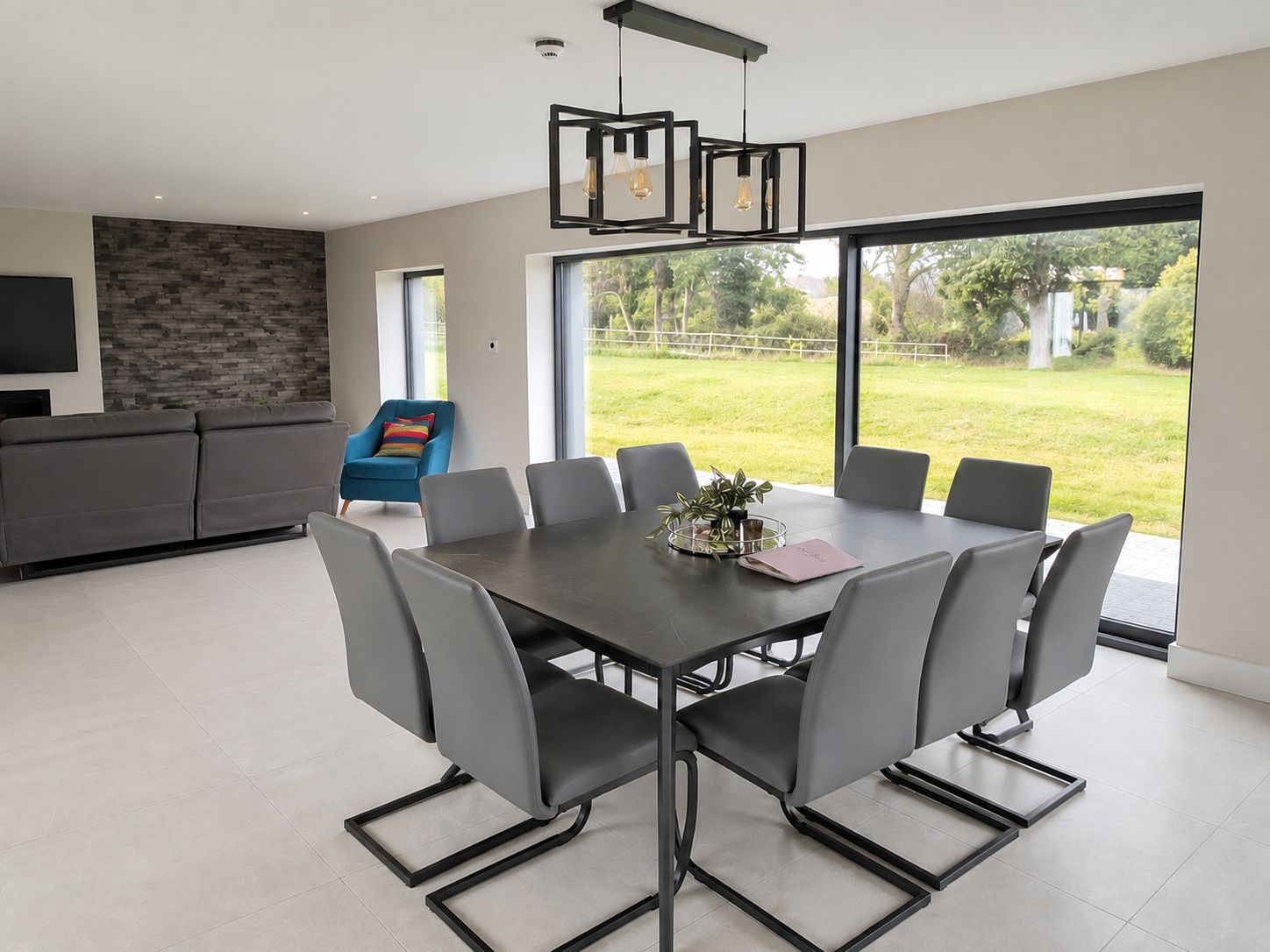 A modern dining area with a large table and chairs, a blue armchair, and a view of a lush garden through large windows.