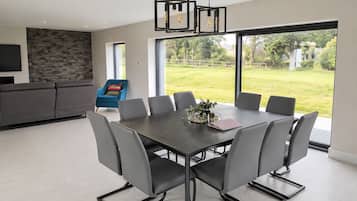 A modern dining area with a large table and chairs, a blue armchair, and a view of a lush garden through large windows.