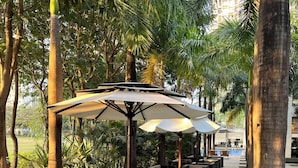 An outdoor seating area with wicker furniture and umbrellas, surrounded by palm trees and a tiled walkway. - Cozy 1-bedroom apartment with AC and golf course access (Gahunje)