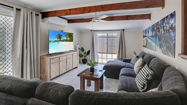 A living room with a grey sofa, a wooden coffee table, a TV on a wooden cabinet, and a large window with a view of a beach. - Fantastic beach house tucked away in paradise (Amity)