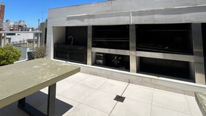 A rooftop terrace with a metal table, a modern building with large windows and a chimney, and a clear blue sky. - Apartment in Nuñez (Buenos Aires)
