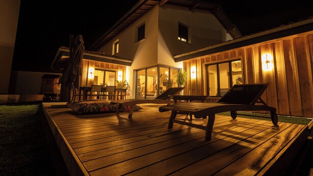 Een houten terras met loungestoelen en een tafel, omgeven door een huis met verlichte ramen en een verzorgd gazon.