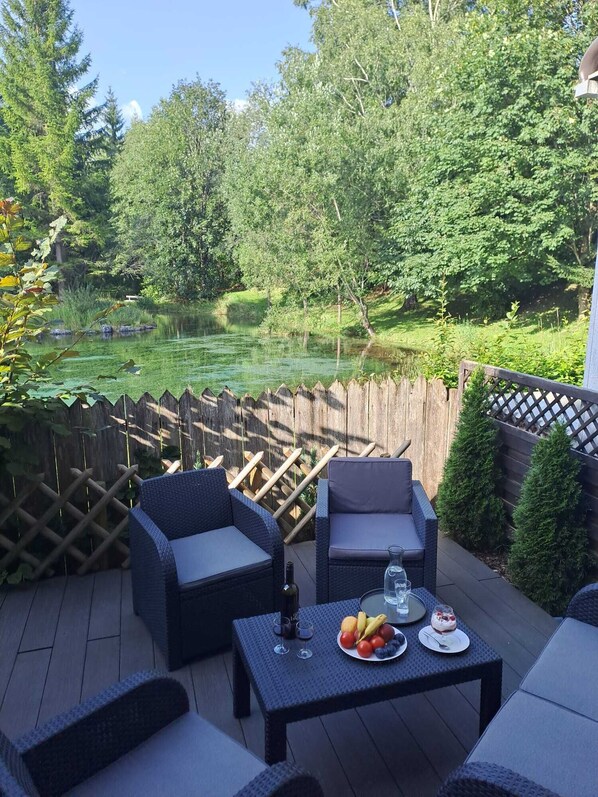 Un patio exterior con mobiliario de mimbre, una mesa con fruta y bebidas, y vistas a un estanque rodeado de árboles.