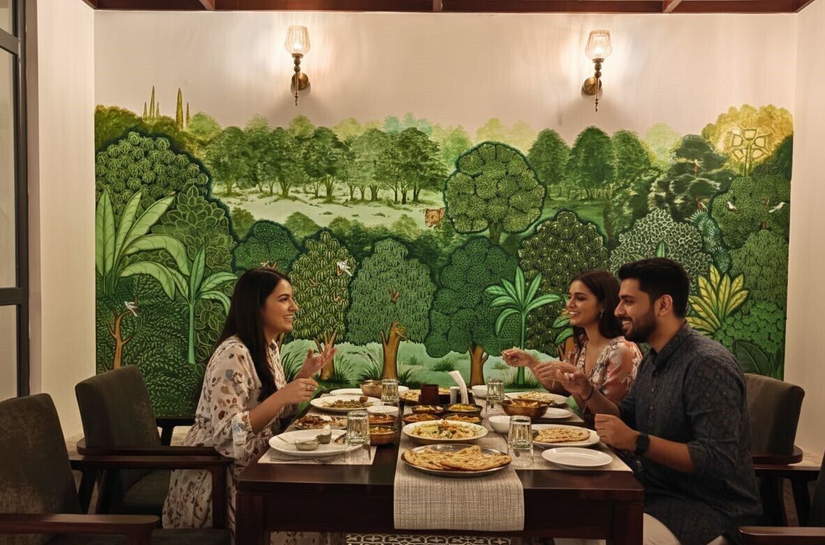 A dining area with a mural of a forest scene, a table set with plates and glasses, and two wall-mounted lamps.