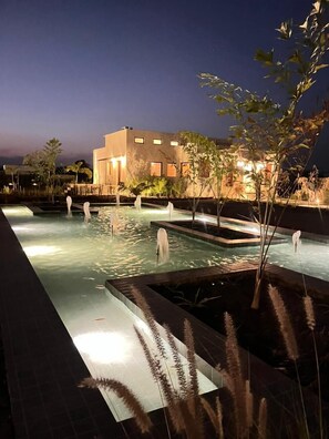 Un espace piscine nocturne doté de plusieurs éléments aquatiques et d’un bâtiment en arrière-plan.