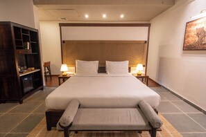 A hotel room with a large bed, two bedside lamps, a wooden headboard, a small cabinet, and a wall-mounted artwork. - Vanthra Vilas (Bali)