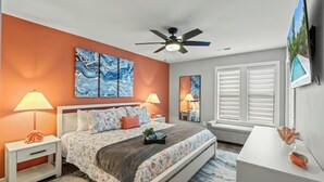 Une chambre à coucher comprenant un lit, deux tables de chevet, un ventilateur de plafond et une grande fenêtre avec des stores.