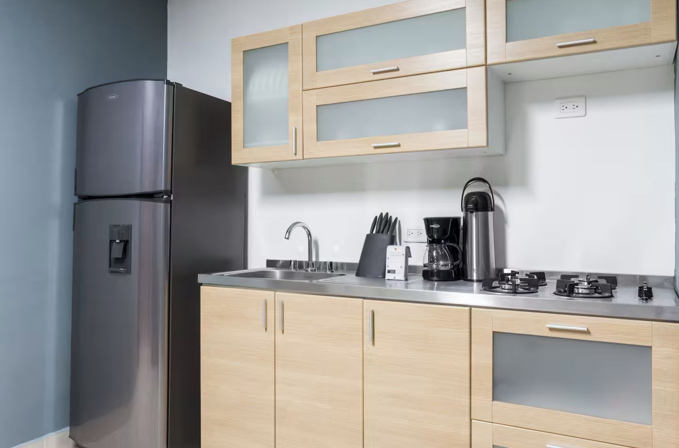 A modern kitchen with a stainless steel refrigerator, wooden cabinets, a coffee maker, and a gas stove.