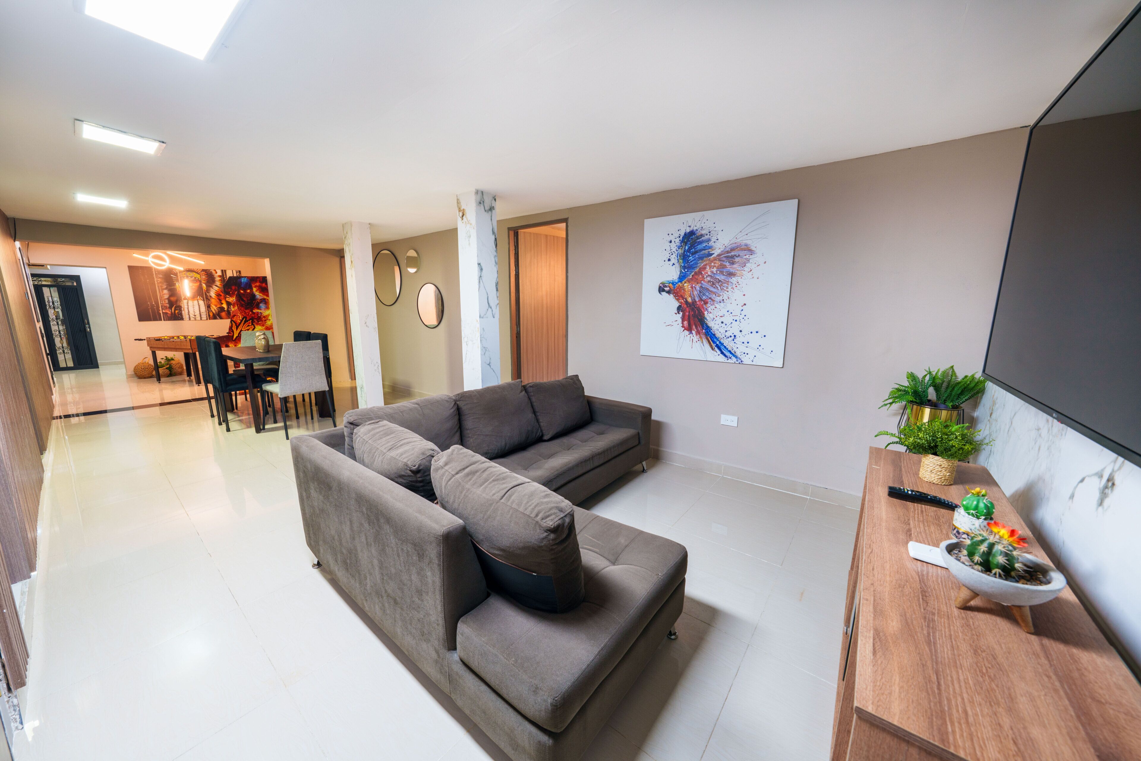 Een woonkamer met een hoekbank, een eethoek met een tafel en stoelen, een wandgemonteerde televisie en een kamerplant.