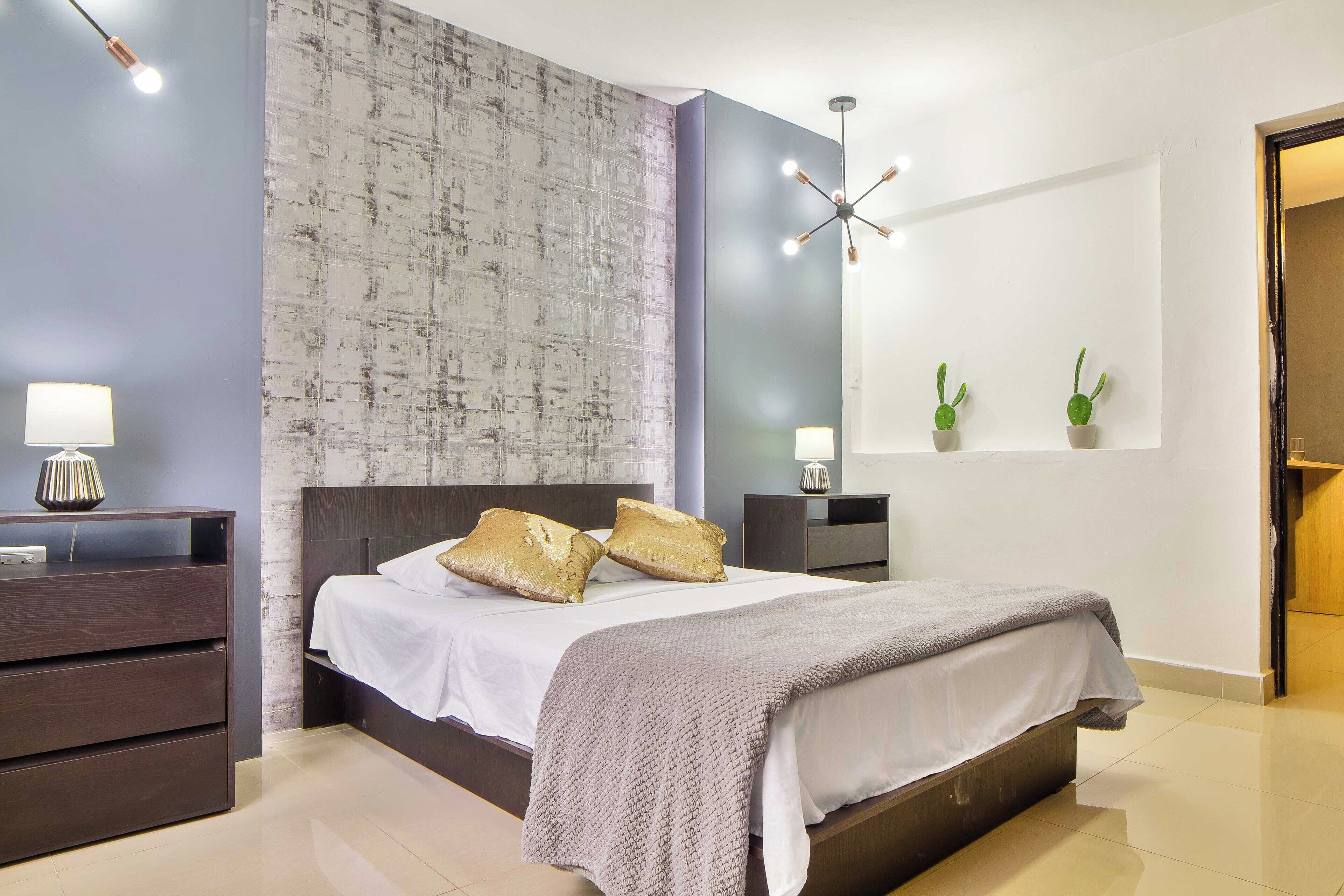 A modern bedroom with a large bed, two bedside tables, a nightstand, a wall-mounted mirror, and a ceiling light fixture.