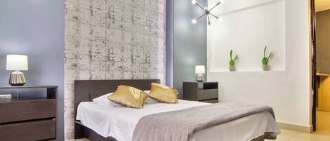 A modern bedroom with a large bed, two bedside tables, a nightstand, a wall-mounted mirror, and a ceiling light fixture.