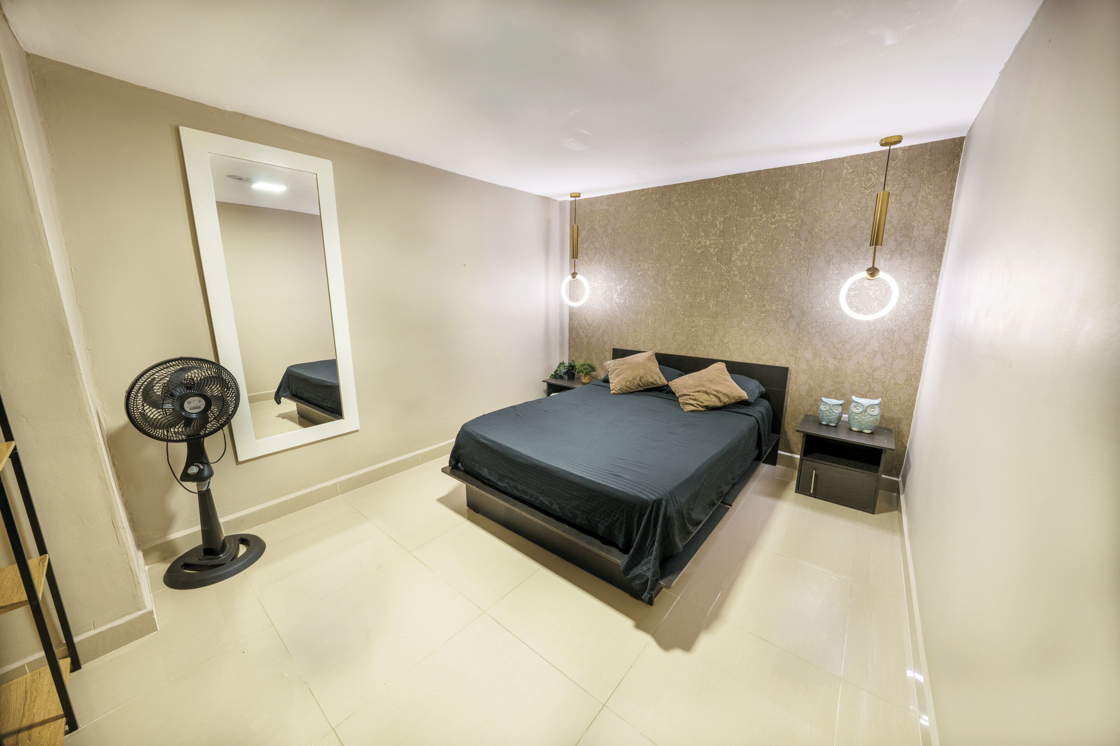 A bedroom with a bed, a standing fan, a nightstand, and a wall-mounted mirror.