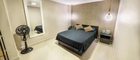 A bedroom with a bed, a standing fan, a nightstand, and a wall-mounted mirror.