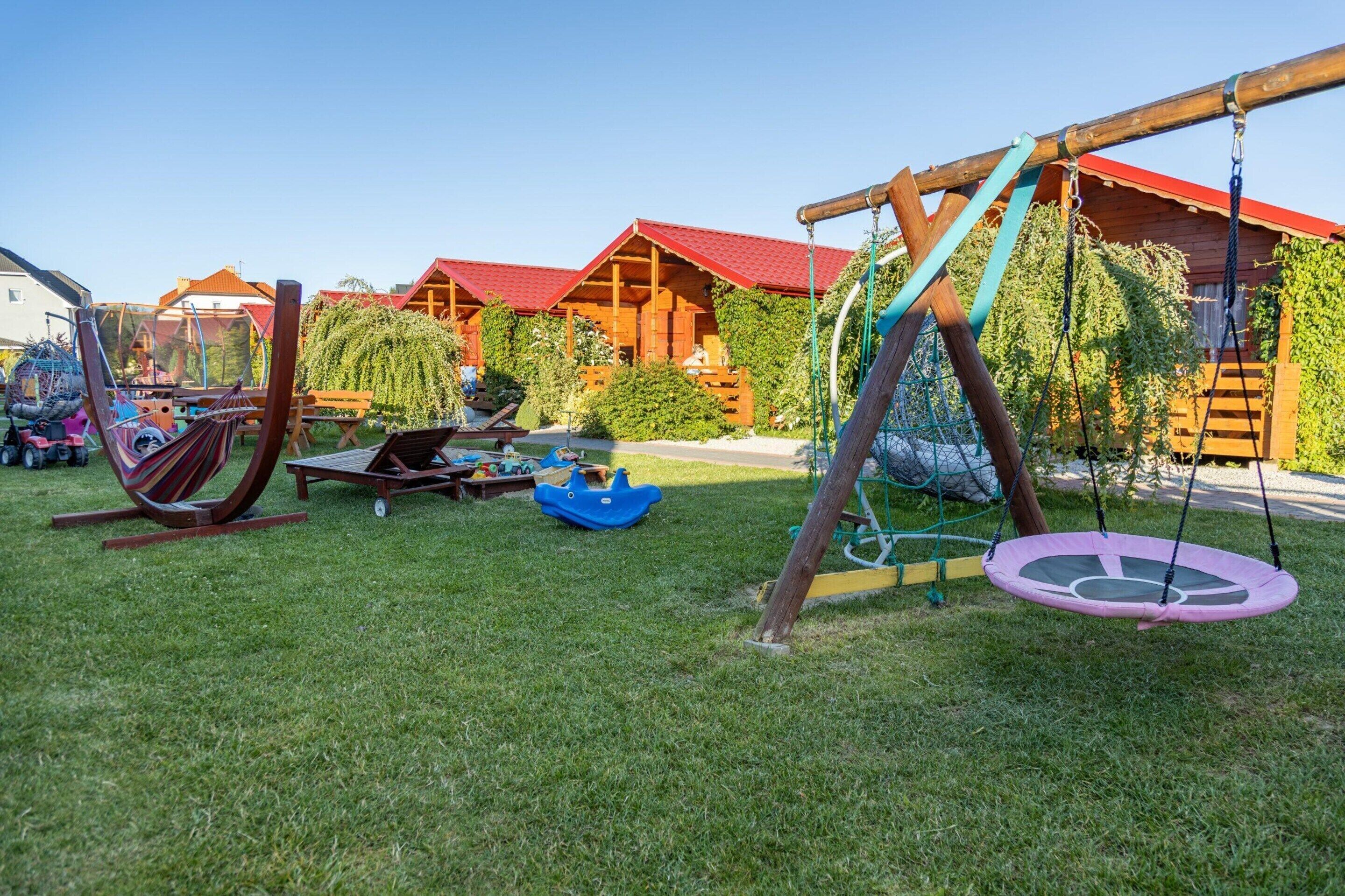 Ein Spielplatz mit Schaukeln und einer Rutsche, umgeben von Holzhütten mit roten Dächern.