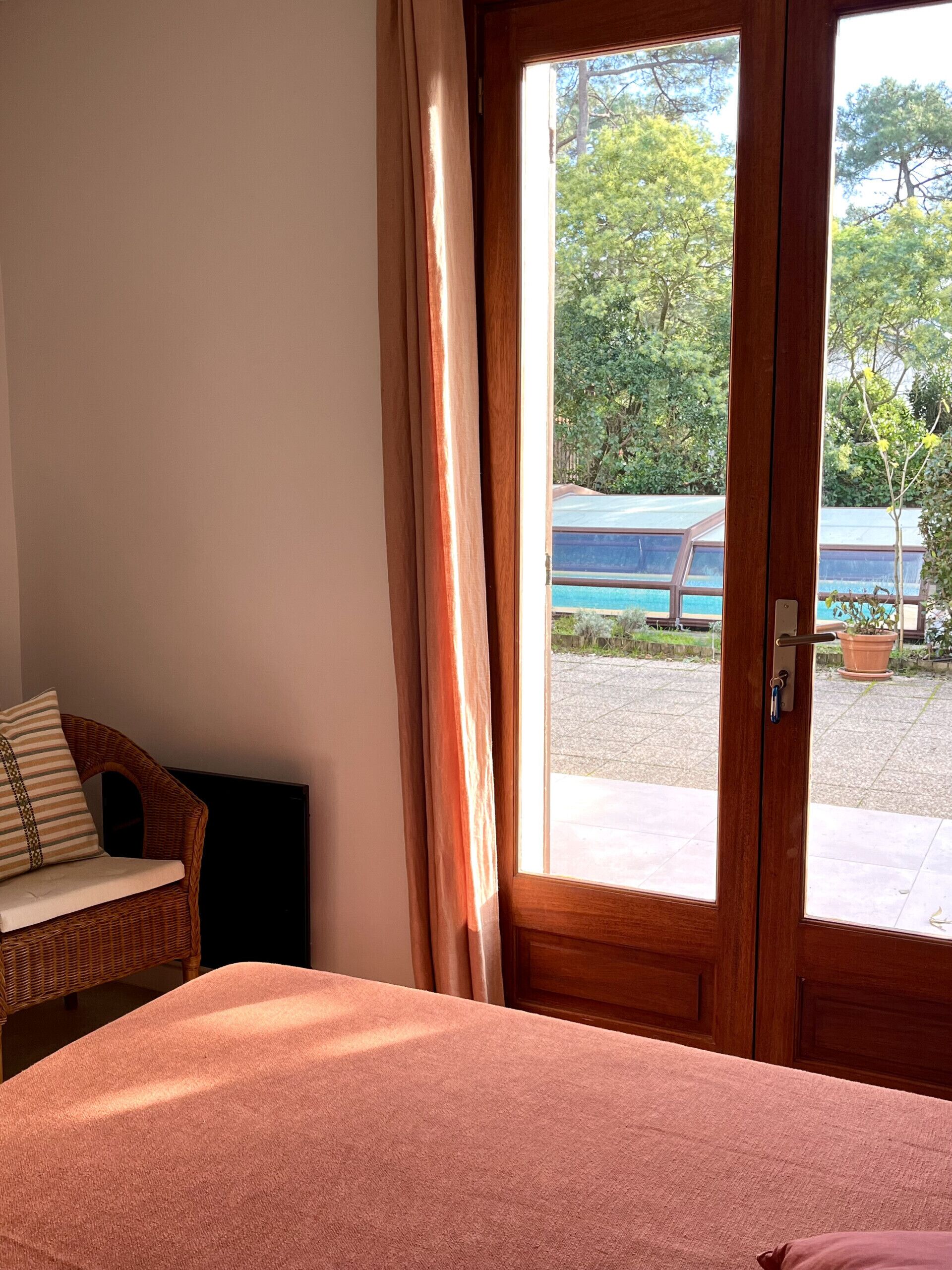 A room with a bed, a wicker chair, and a view of a pool and greenery outside.