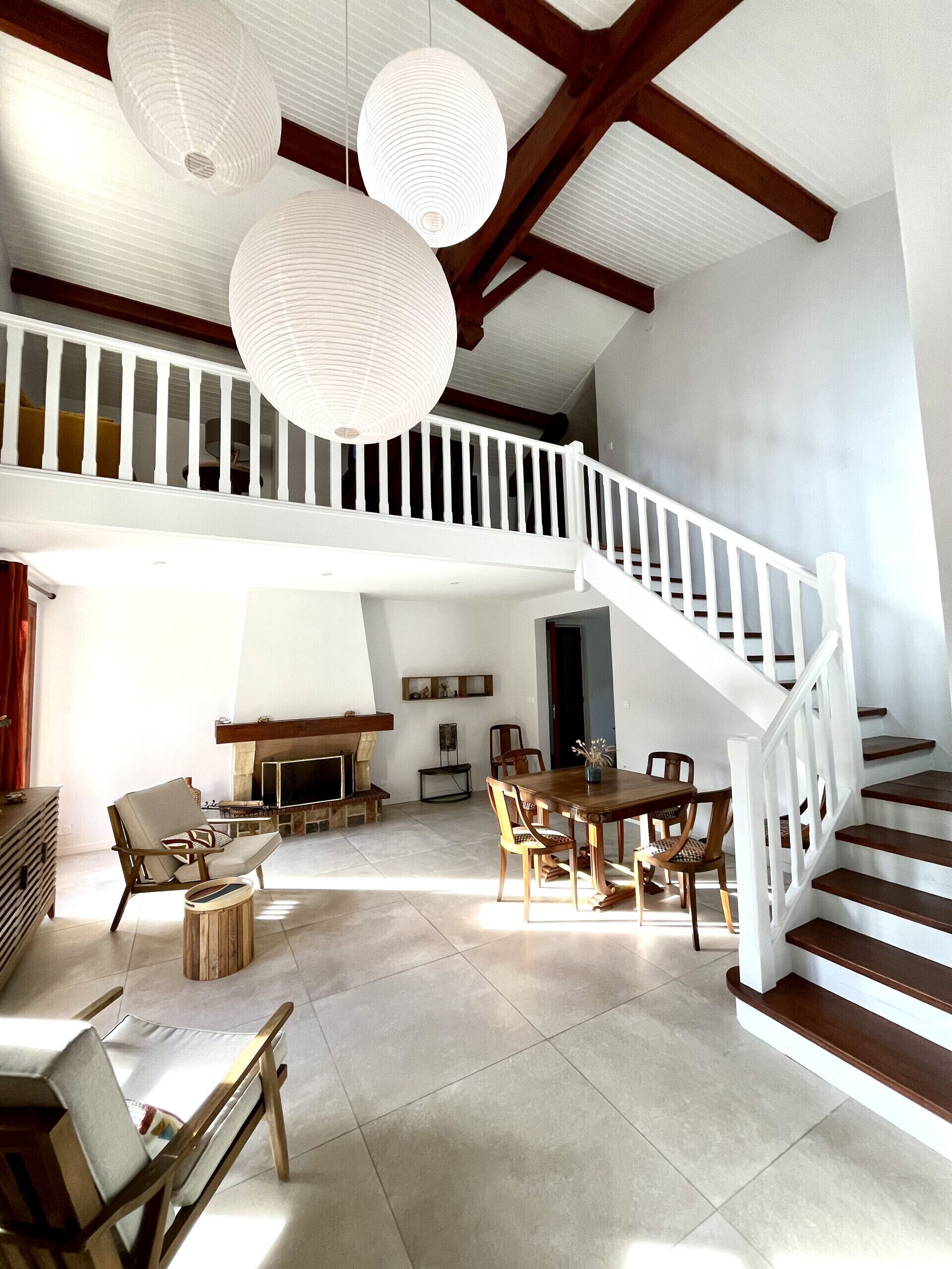 A spacious living area with a high ceiling, wooden beams, and a white staircase. There are several chairs and a dining table.
