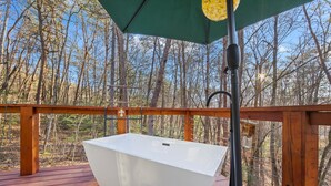 Un évier extérieur blanc, installé sur une terrasse en bois, est surmonté d’un parasol vert et entouré d’arbres.