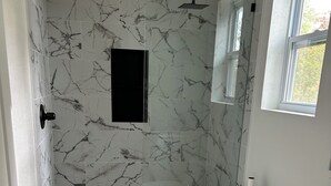 A bathroom with marble-patterned walls, a white toilet, a shower with a handheld showerhead, and a window providing natural light. - Welcome to the Savanna Shire! Cozy one room cabin for a lovers getaway! (Thomson)