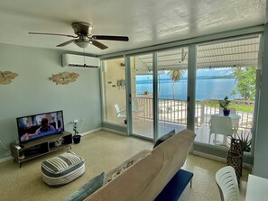 Een woonkamer met een televisie, een plafondventilator en een schuifdeur naar een balkon met uitzicht op de zee.