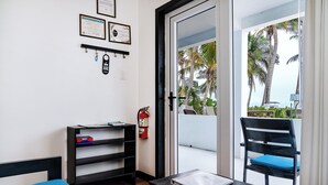 Una habitación con una cama azul, un escritorio negro con un libro, una silla, un extintor y vistas a palmeras a través de una puerta de cristal.