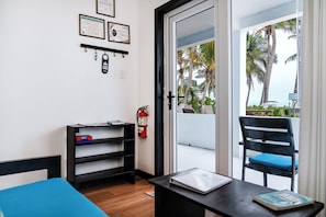 A room with a blue bed, a black desk with a book, a chair, a fire extinguisher, and a view of palm trees through a glass door. - Beachfront Corner Suite | Ground Floor Ocean Views – Aqua Vista Suite 1 (San Pedro)