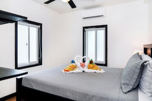 A hotel room with a bed, two windows, a ceiling fan, and an air conditioning unit. - Beachfront Corner Suite | Ground Floor Ocean Views – Aqua Vista Suite 1 (San Pedro)