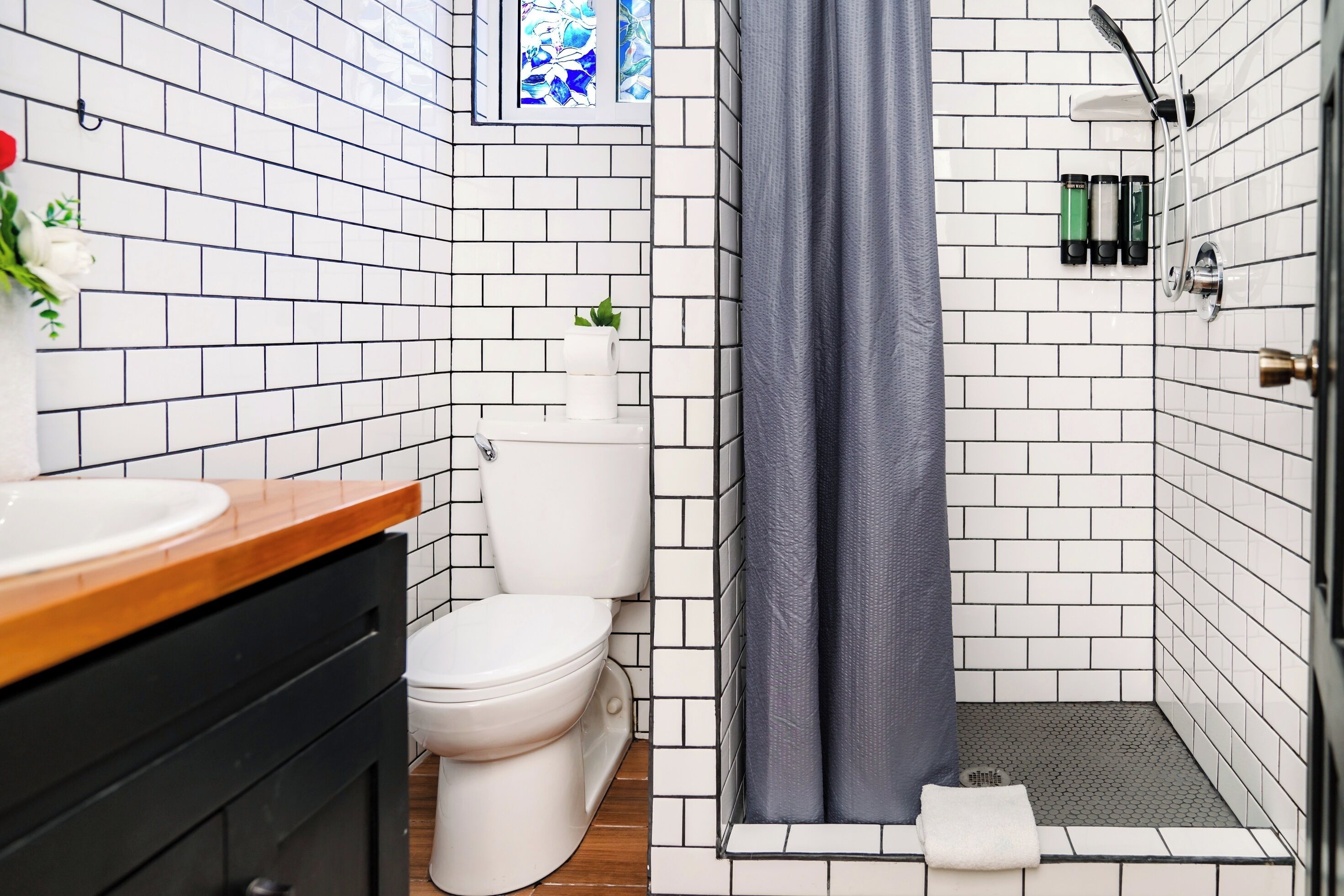 Une salle de bain avec des murs recouverts de carreaux blancs, un plan de toilette en bois, des toilettes blanches, une douche avec un rideau gris et une petite fenêtre ornée de motifs bleus et blancs.