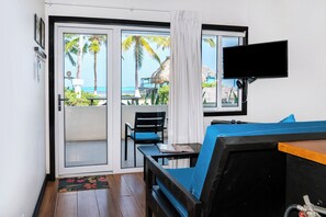 Habitación con puerta corrediza de vidrio que da a un balcón con vistas a palmeras y al océano. Se ve un televisor montado en la pared, una mesa con una silla y un sofá azul.