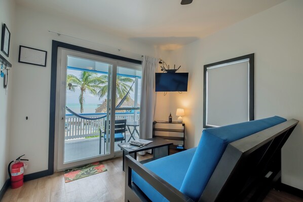 Een kamer met een blauwe bank, een televisie, een tafel met een lamp, en uitzicht op het strand door een schuifdeur.