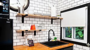 Una cocina con un refrigerador negro, una cafetera, un lavamanos y un ventanal con follaje verde en el exterior.
