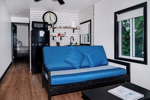 A compact living space with a sofa, a small table, a microwave, a refrigerator, a clock, and a window with a view of greenery. - Top-Floor Corner Beachfront Suite with Reef Views | Aqua Vista – Suite 6 (San Pedro)