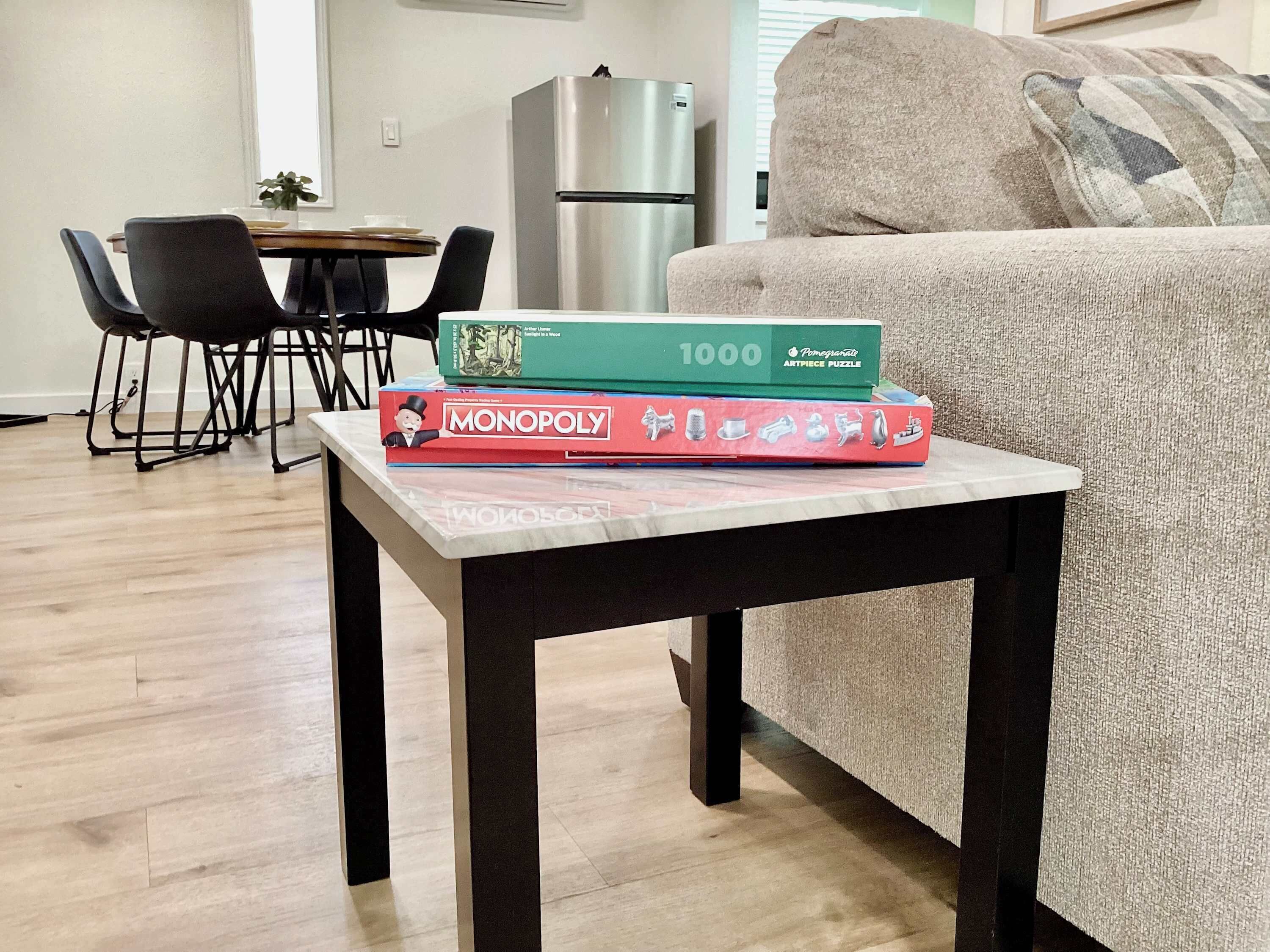 A living room with a table holding board games, a sofa, a dining area with chairs, and a refrigerator.
