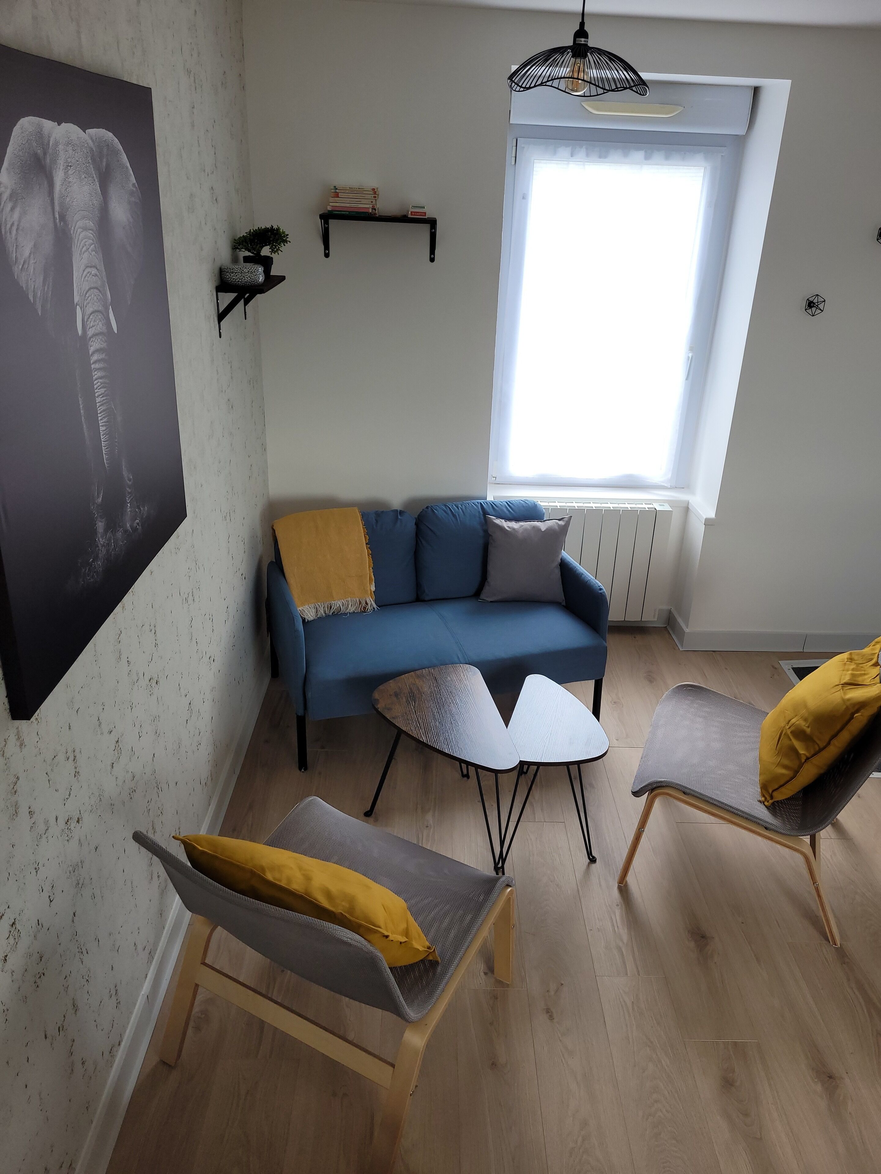 A living room with a blue sofa, two chairs, a small table, a large window, and a wall-mounted shelf with books.