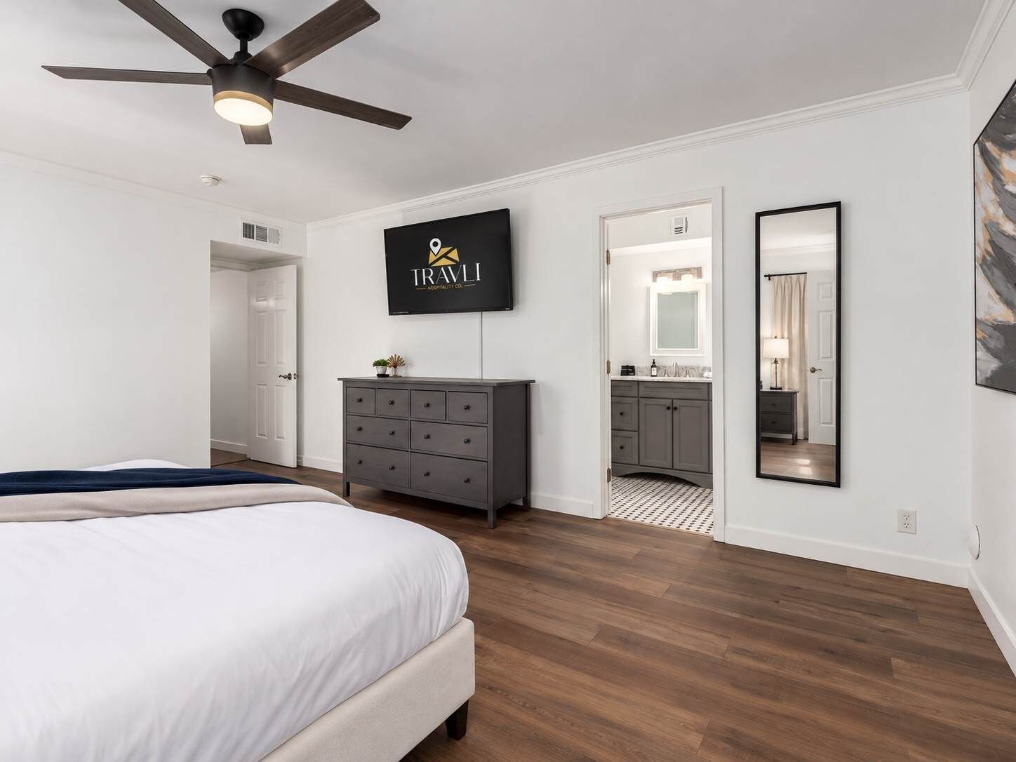A bedroom with a bed, a dresser, a ceiling fan, a TV, and a bathroom with a mirror and sink.