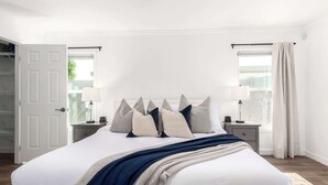 A bedroom with a large bed, two bedside tables with lamps, a door, and windows with curtains. - The Desert Bloomhouse | Heated Pool |Sleeps 12 (Scottsdale)