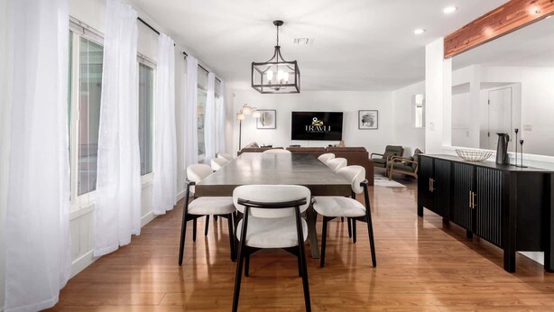 A modern dining area with a long table, white chairs, a black cabinet, and a television on the wall. - The Desert Bloomhouse | Heated Pool |Sleeps 12 (Scottsdale)