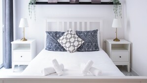Un lit bien fait, avec du linge de lit blanc et deux oreillers, flanqué de deux tables de chevet avec des lampes, dans une chambre dotée d’une tête de lit blanche et d’une étagère au-dessus du lit.