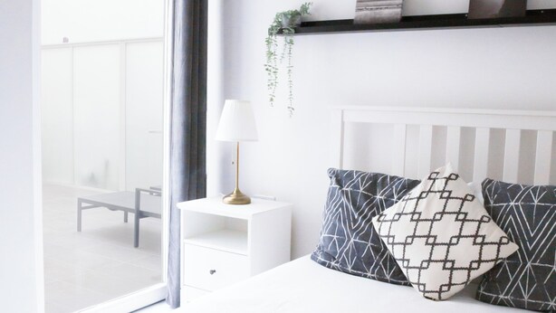 Une chambre avec une grande fenêtre, un lit recouvert de draps blancs, une table de chevet avec une lampe et un climatiseur fixé au mur.