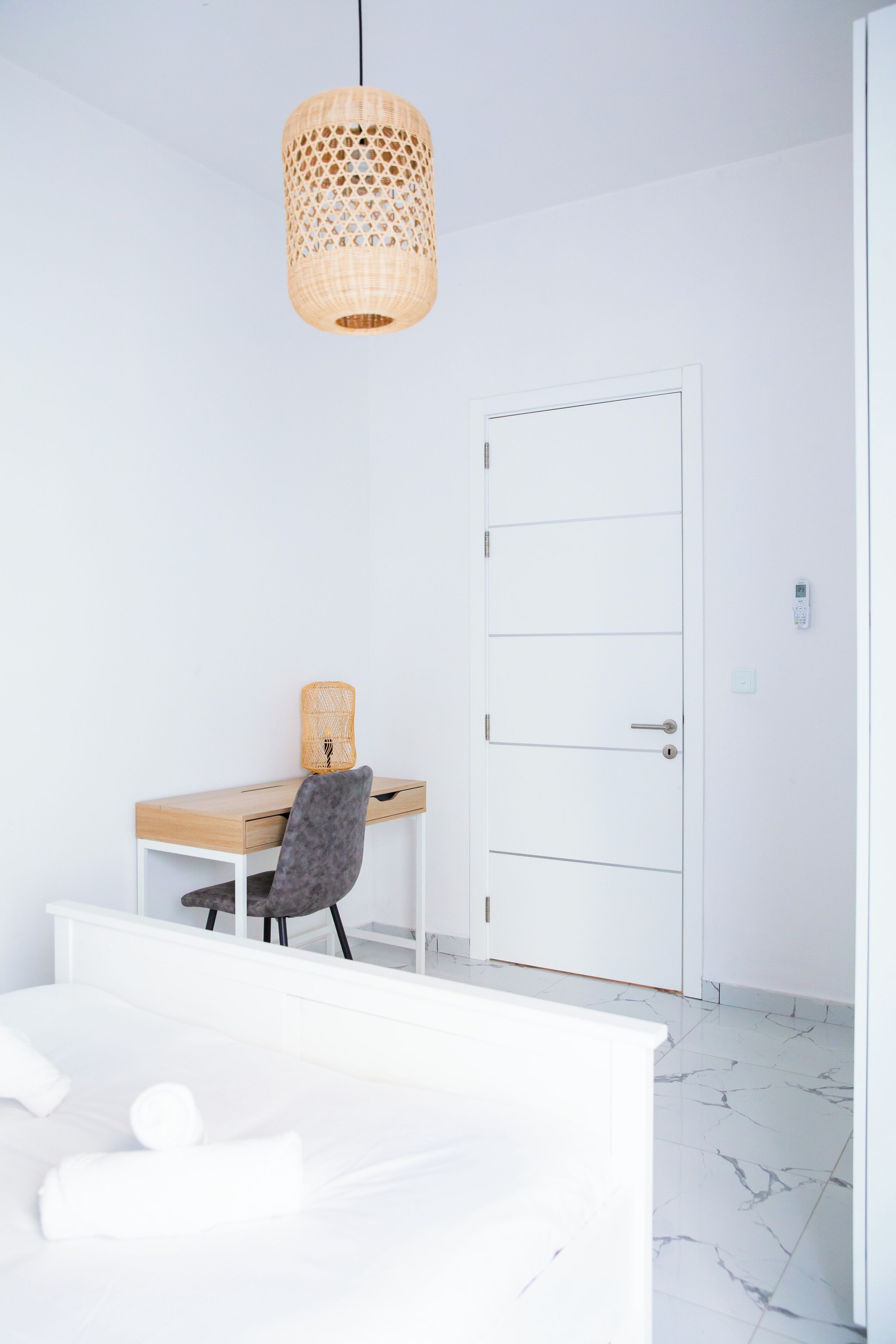 Ein modernes, minimalistisches Zimmer mit einem Holztisch, einem grauen Sessel, einem weißen Bett und einer weißen Tür.