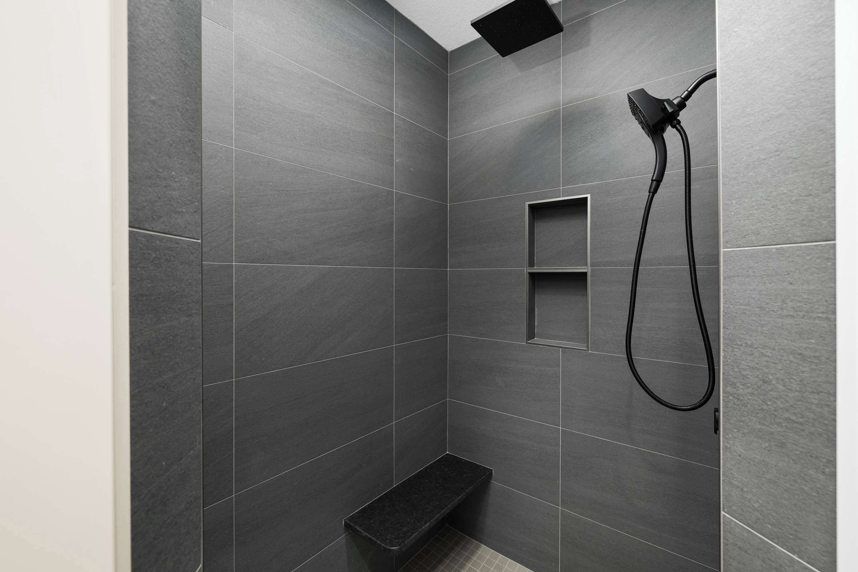 Un espace douche moderne avec des carreaux de couleur sombre, une douchette à main et une petite étagère.