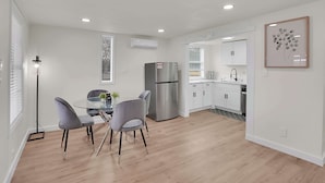 A modern kitchen with white cabinets, a stainless steel refrigerator, and a dining area with a glass table and grey chairs. - Boardwalk Bungalow 1 For 6 (League City)