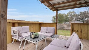 An outdoor patio with a wooden deck, white cushioned seating, and a glass table. - Boardwalk Bungalow 1 For 6 (League City)