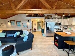 Ein gemütliches Wohnzimmer mit einer Couch und einem Hocker, eine Küche mit Kühlschrank und Mikrowelle sowie eine Holzbalkendecke.