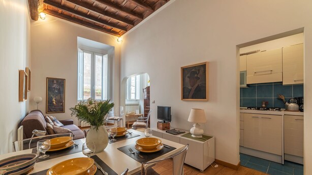 Uma área de refeições com uma mesa posta para quatro pessoas, um sofá, uma TV e uma cozinha visível ao fundo.