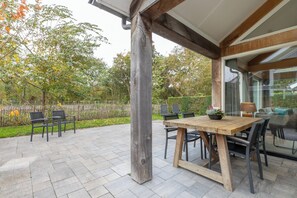 Una veranda esterna con tavolo e sedie in legno, pavimentata con pietra, e vista su un giardino con alberi e una recinzione.