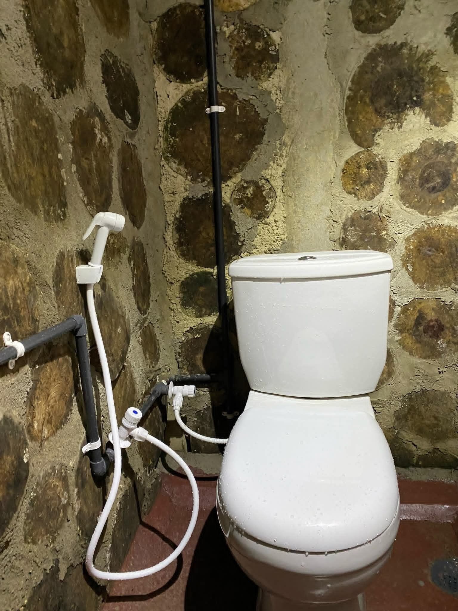 Sanitário branco com cano preto e cano branco fixados, com parede de pedra ao fundo.