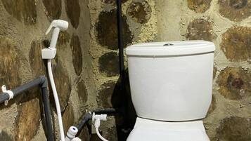 Sanitário branco com cano preto e cano branco fixados, com parede de pedra ao fundo.